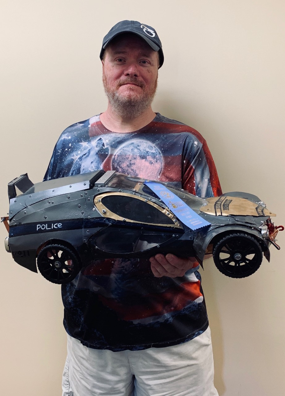 Photo of Brent, a man with disabilities, is wearing a baseball cap and T-shirt and holding a very large model car in his hands