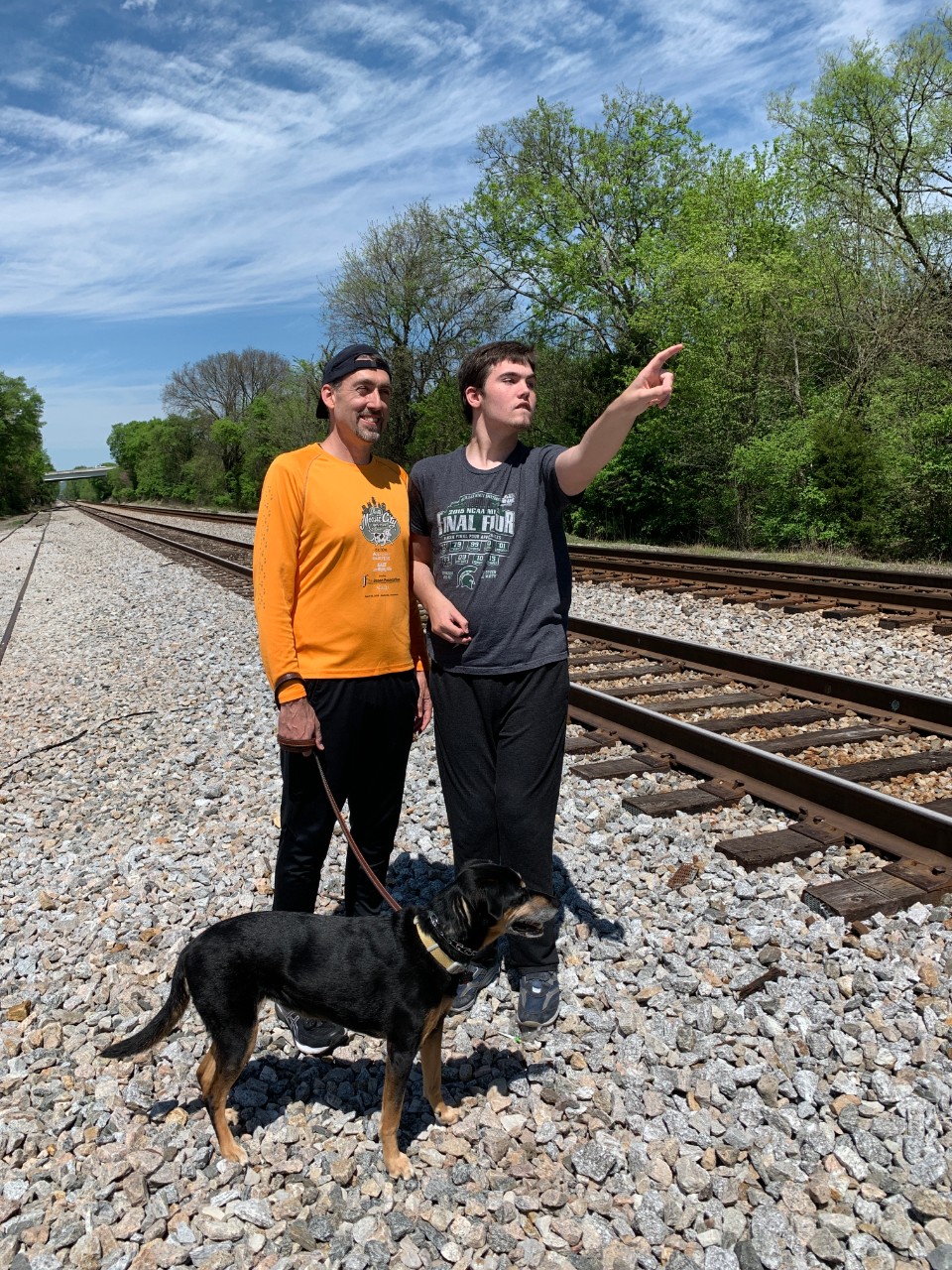 A photo with Aidan and his father standing next to train tracks on a beautiful sunny day, and Aidan pointing to a train past the camera. They're also walking their medium sized black dog.