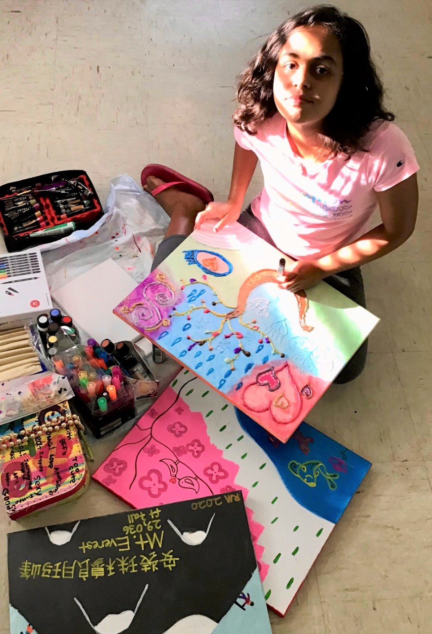 photo of the young artist – she is shown sitting on the floor with several paintings of hers around her and in her lap, doing her art. There’s sun from a window shining on her face, and she has a hint of a smile