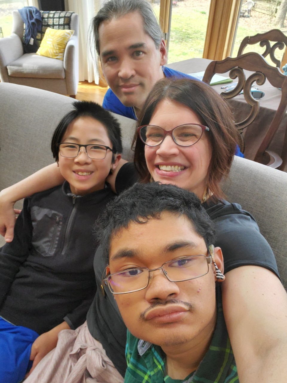 a family photo of the Guilarans – husband and father Fonsie is kneeling behind a couch where the rest of the family is sitting; the younger son Xiao Yu on one side of mom and wife Leslie, and the older teen son Angel on the other side; Leslie’s arms are around both of her sons. Leslie is a white woman with short brown hair, glasses and a big smile. Both sons are Southeast Asian and have short black hair and wire-rimmed glasses. Angel has a short mustache and a visible cochlear implant on one ear. Fonsie is Filipino and has long black hair pulled back in a ponytail
