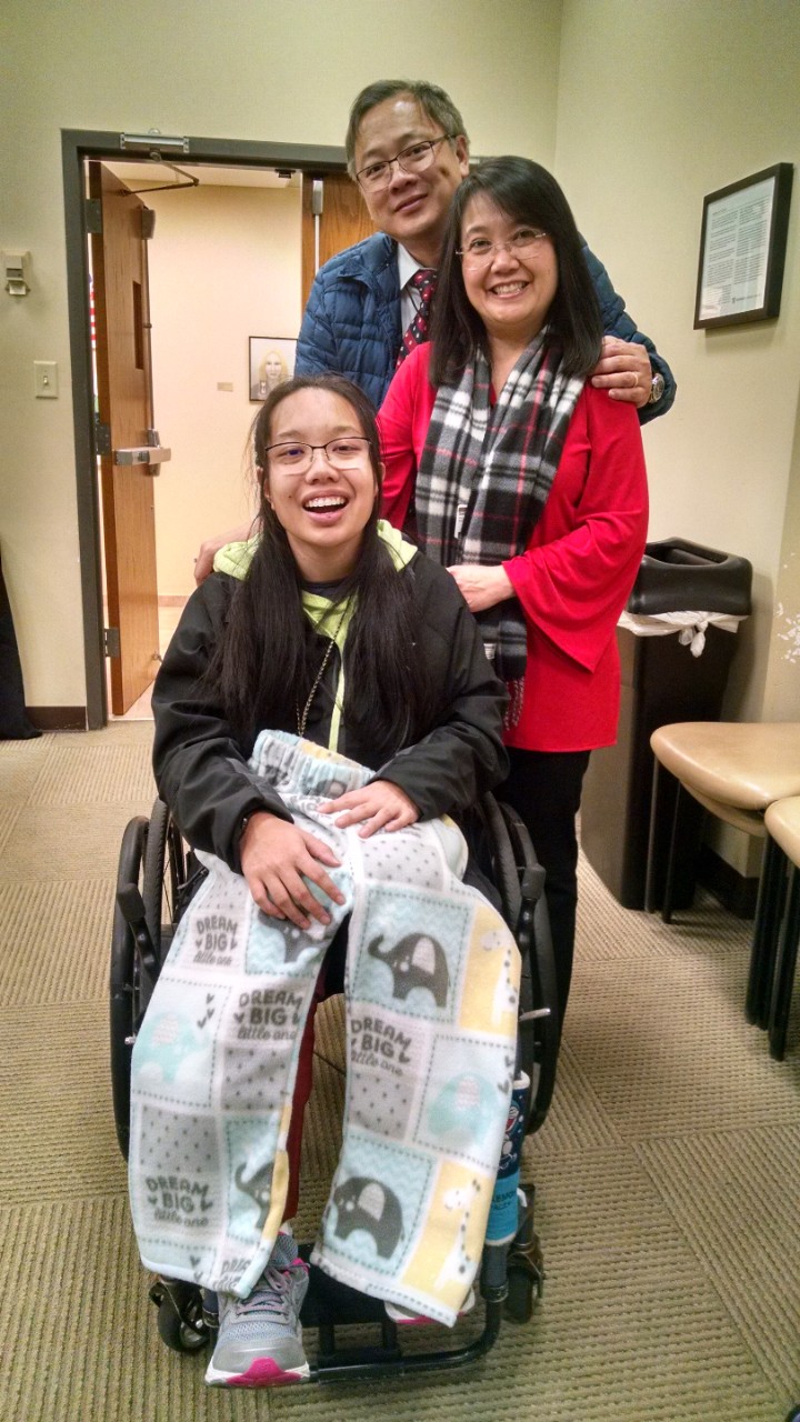 A young Asian woman with long black hair, glasses and a smile sits in her wheelchair with her parents standing behind her. She’s holding soft fleece patterned pants across her legs.