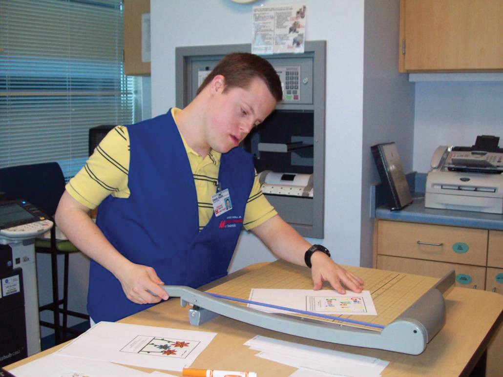 Edward Nesbitt when he was at Next Steps, working at one of his internships. He is wearing a blue worker vest and using a cutter to trim some paper.