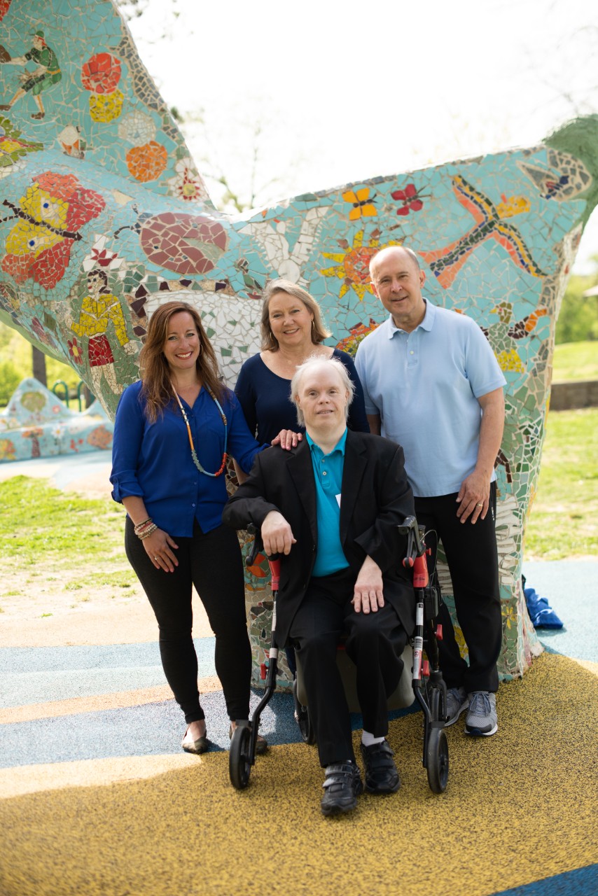 Photo shows Scott, an older gentleman with Down syndrome with white hair seated on a walker, wearing a nice black suit with a blue collared shirt. Behind him stand his supporters, mentioned in the caption. All are smiling, in front of a blue mosaic statue or mural with pretty insect and flower designs.