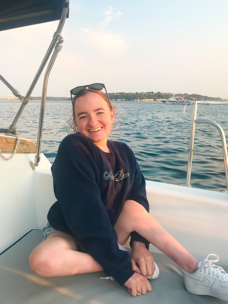 Rosie took a sailing excursion in Seattle, Washington. She is shown sitting on the bench of a boat, smiling with the ocean behind her. She is in a navy sweatshirt, has her hair pulled back into a ponytail, is wearing sunglasses on the top of her head, and shorts with white sneakers.