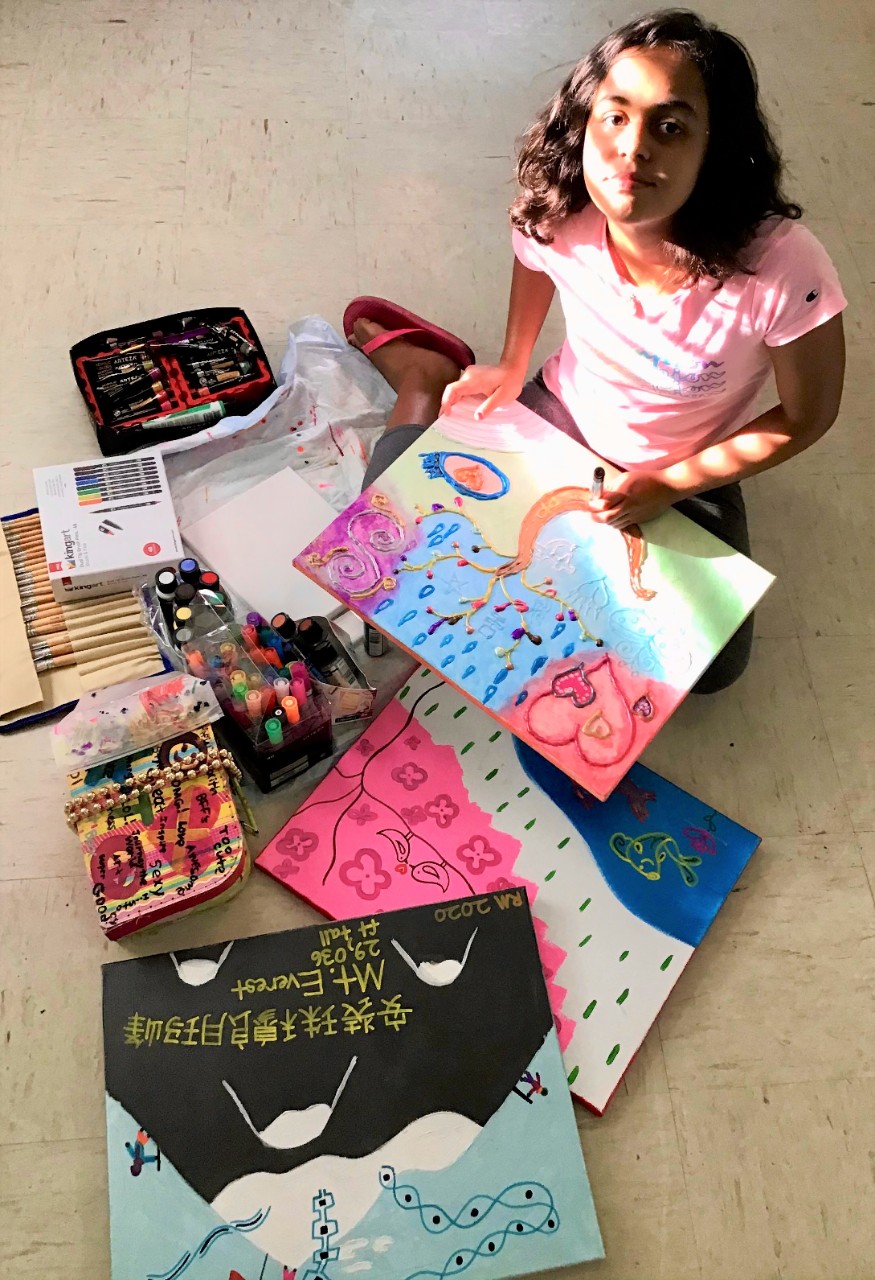 A young girl with dark skin and dark, wavy hair looks up from her seated position on the floor, surrounded by paintings and art supplies. Slatted sunlight shines on her face and pink t-shirt
