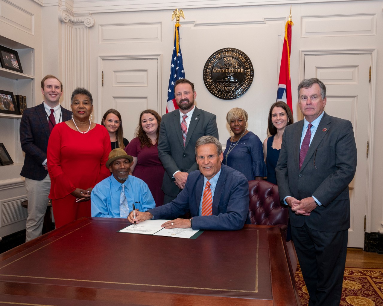 A group of 10 people pose in a formal meeting room with the Tennessee state seal and the U.S. and Tennessee flags in the background. Governor Lee is seated at a table, with a document and pen in front of him. Council member Martez Williams, a middle-aged Black man who uses a wheelchair, is sitting to the Governor’s right at the table. Behind them is a group of adults, both men and women, Black and white, wearing business attire