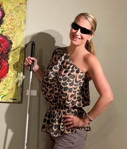 1.	Photo of Tracy shows her posing beside one of her painting in a gallery and smiling with her hand on her hip. She is a white woman with long blonde hair pulled back in a ponytail wearing dark sunglasses, a black and gold patterned blouse that reveals one shoulder, and holding her white cane beside her.