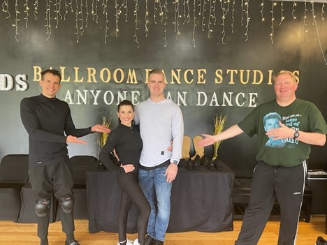two dance students, Ben and Dave, holding their hands out to present their two dance teachers at the center; all are wearing athletic dancewear and the wall behind them reads “Ballroom Dance Studios – anyone can dance.”