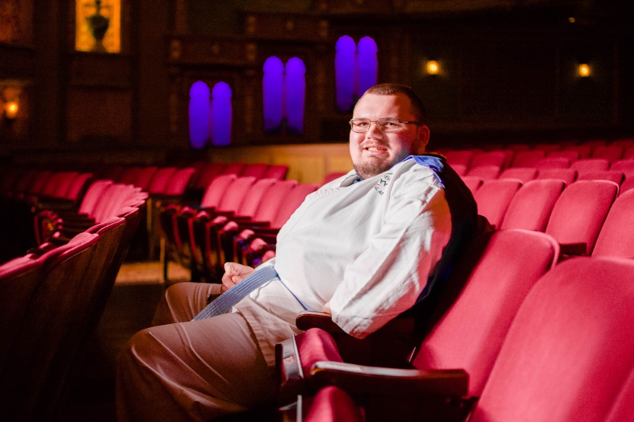 young white man sitting in red velvet seats in an empty theater; wearing glasses and a blue cape