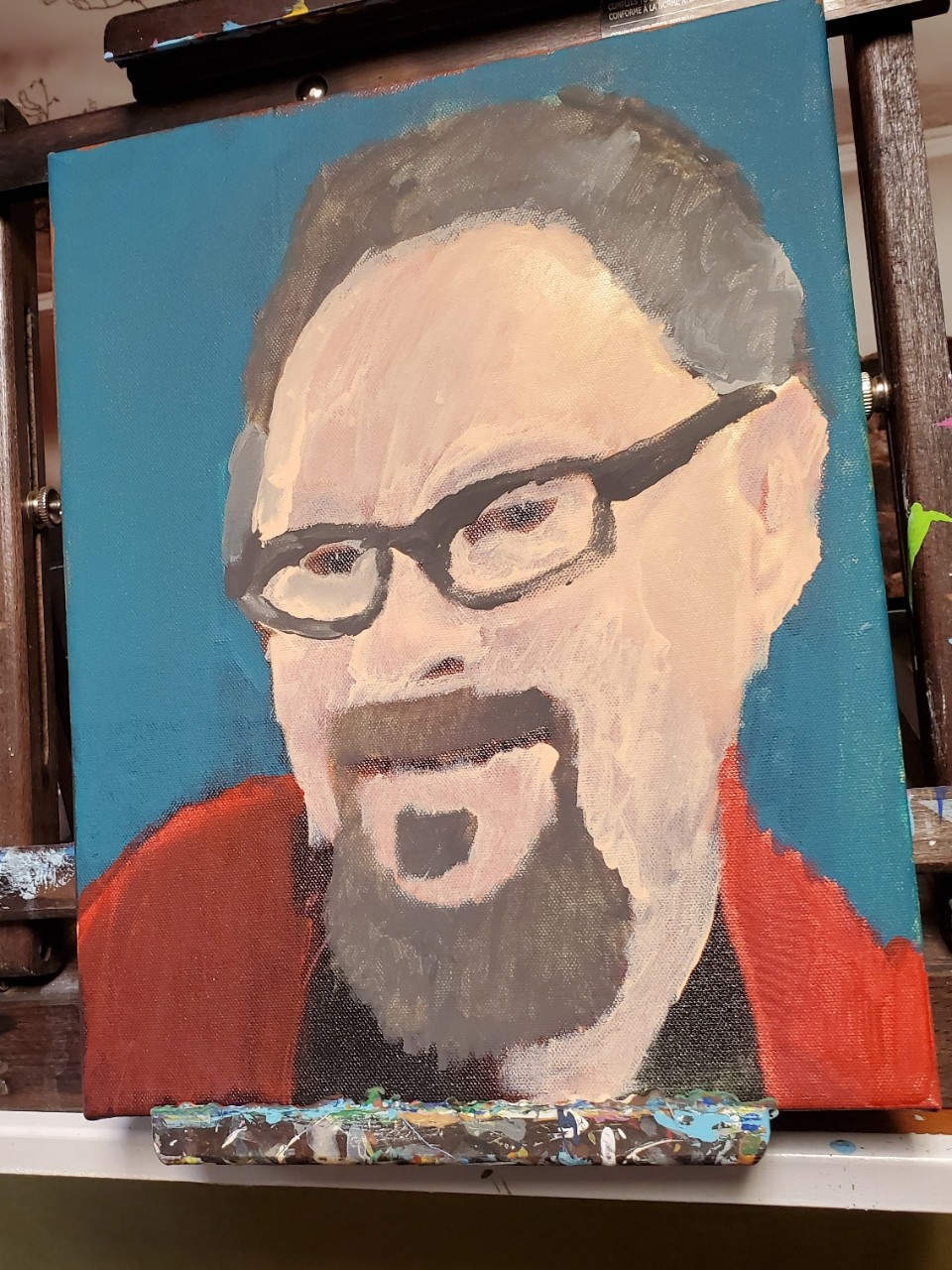 photo of a painting in a studio. The painting is of an unidentified man with brown hair and a brown goatee, glasses and a red shirt, against a blue background.