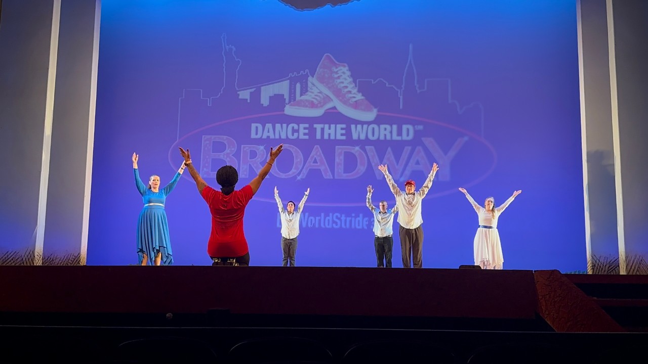 Image description 1: Five adult dancers with disabilities stand on a Broadway stage with their hands raised in identical poses in the air; the 3 men are wearing slacks and button down white collared shirts; the two women are wearing similar flowing belted dresses, one blue and one white. Their instructor and choreographer has her back to the camera and is kneeling in front of the stage facing them. Behind the dancers, it shows a logo for “Dance the World: Broadway” which is a New York skyline and a pair of red sneakers.