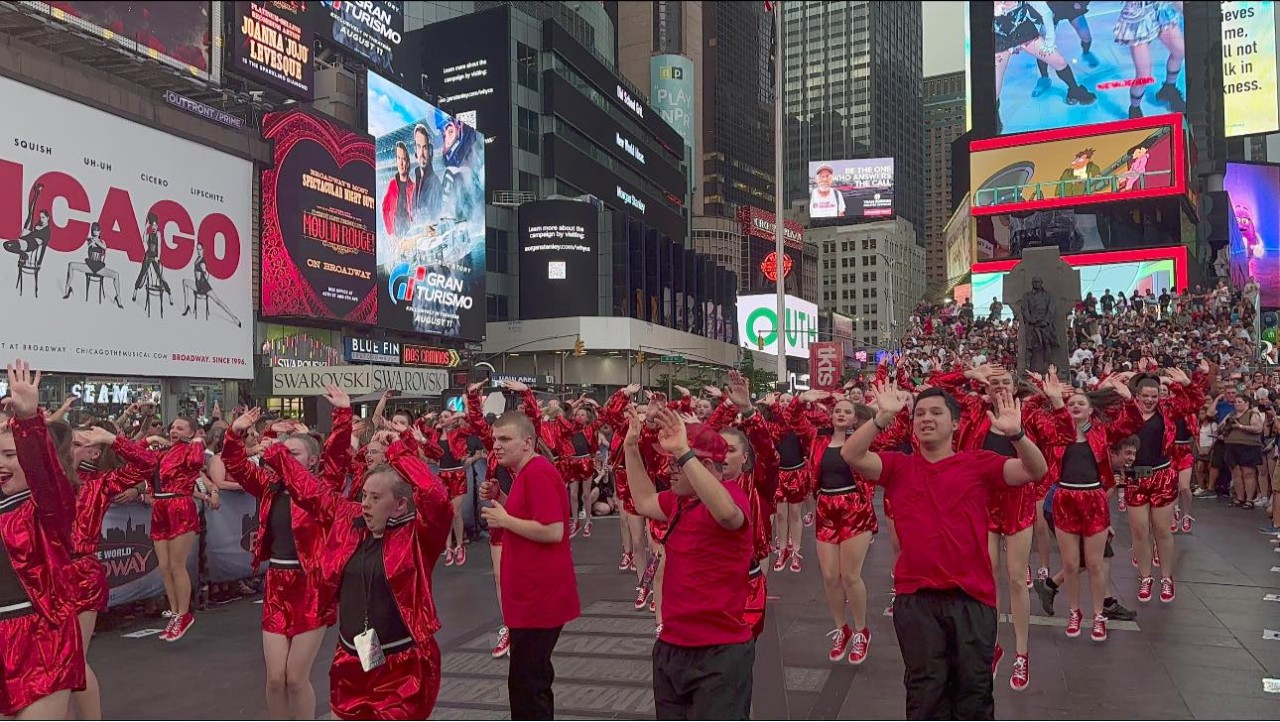 A large group of people are shown doing a choreographed dance down the middle of a large New York city street, all wearing bright red outfits.