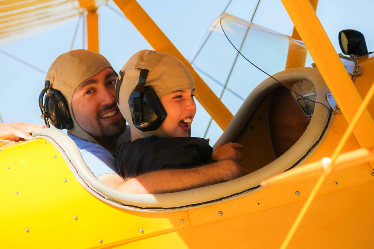  a boy and an older man in the cockpit of a yellow plane. Both males are smiling happily.