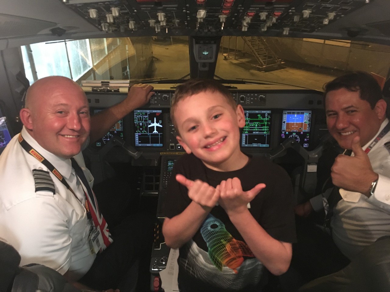 the cockpit of a commercial airplane. There are two pilots smiling at the camera; one is giving a “thumbs up” sign. In the middle is a young boy, Jessica Gant’s son. He is smiling and giving two “thumbs up”. 