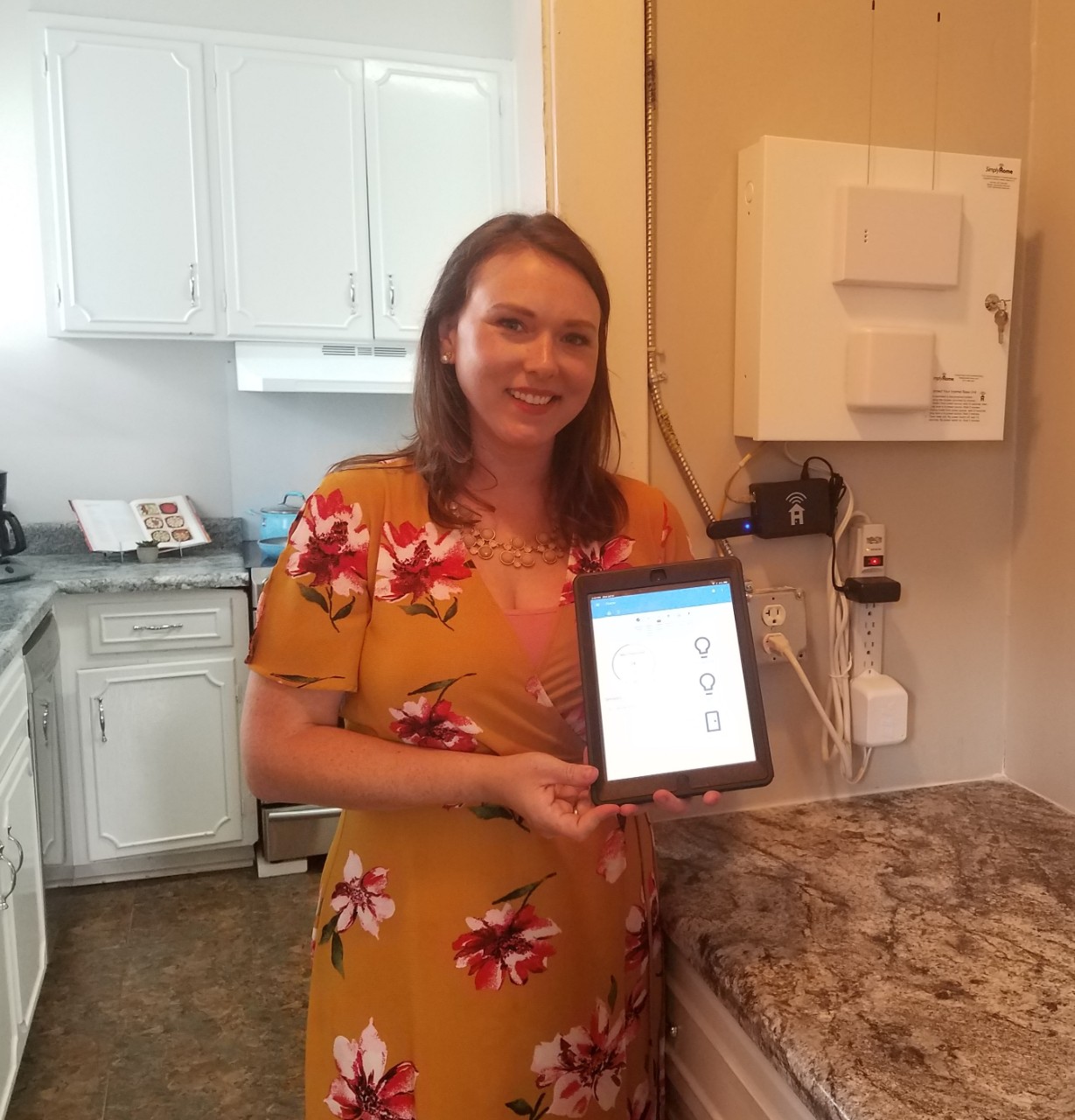 Kate Adams, a young woman in a gold floral dress, stands in the kitchen of the technology model home described in this article and holds up an ipad showing an open app