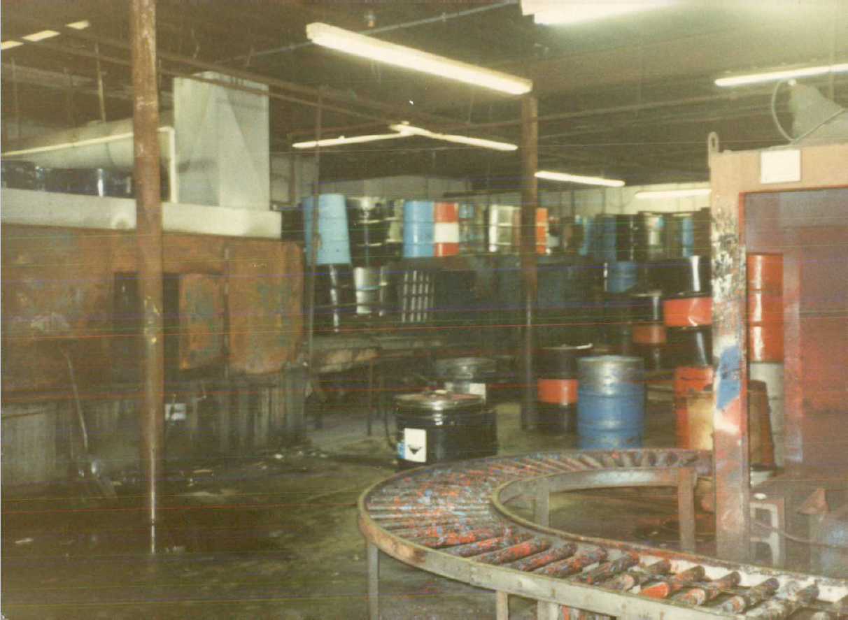 An old photo of the paint-stained conveyor belt inside the Dixie Barrel & Drum facility. Cleaned and restored drums sit on shelves and the floor in the background, and puddles on the concrete slab floor reflect rusty iron beams supporting the building. 