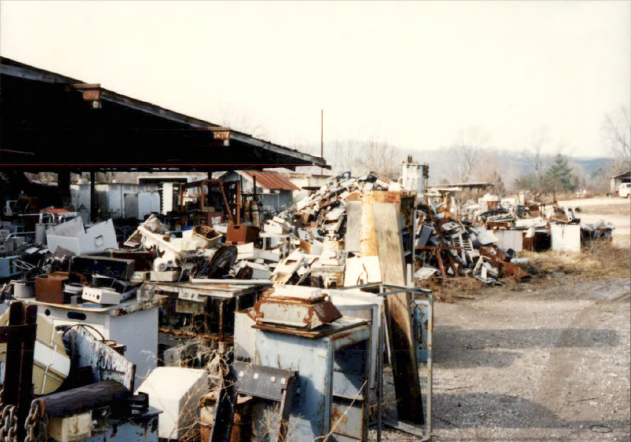 Joyner scrap yard including heaps of miscellany on the bare ground. Items include rusted metal parts, large boxes,  and indistinguishable materials.