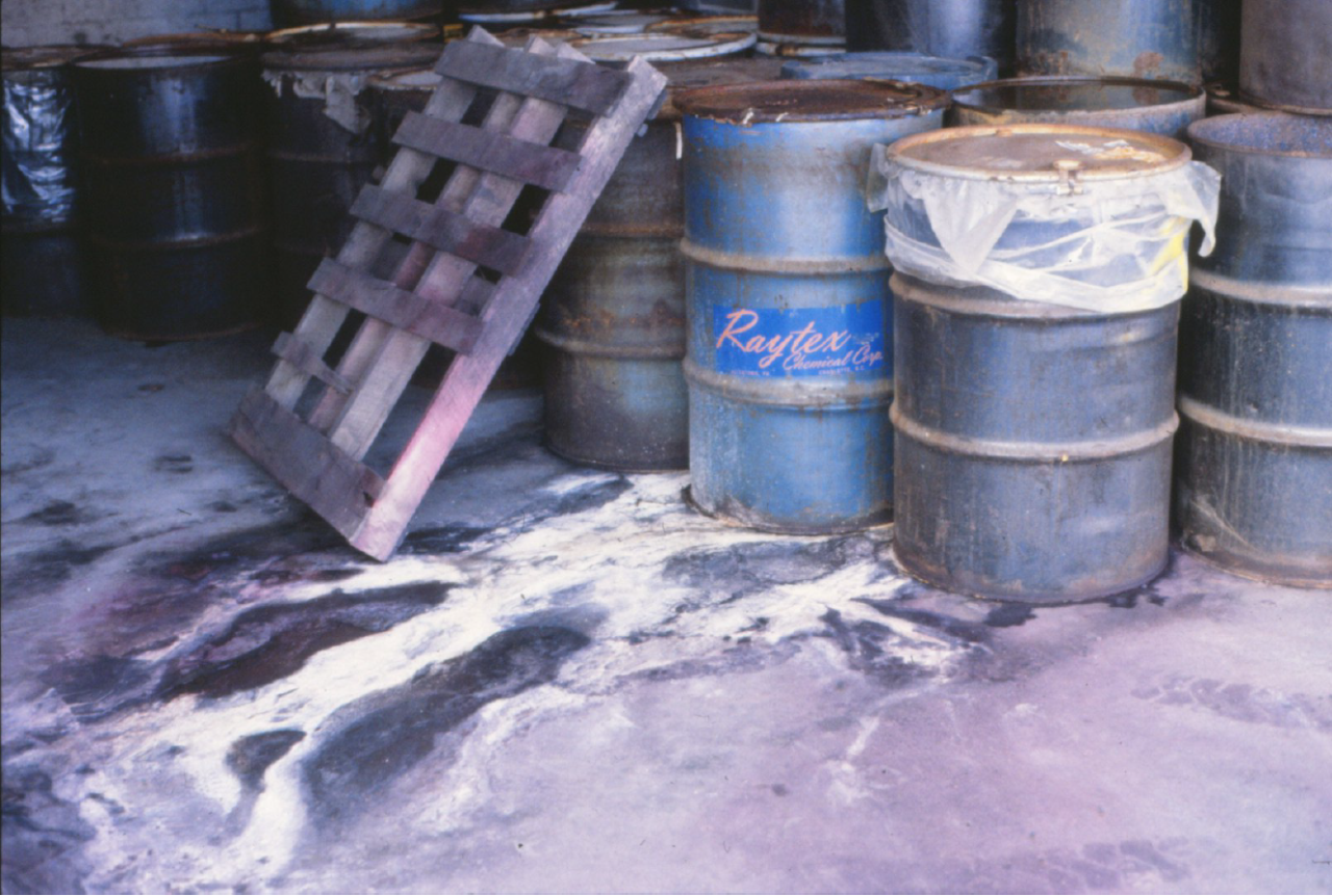 Leaking drums in the Morningside Chemical facility. A wooden pallet is leaned against a dozen drums, mostly gray and rusted with a blue drum labelled "Raytex Chemical" in the foreground that appears to be leaking a white oxide material all over the concrete floor. 