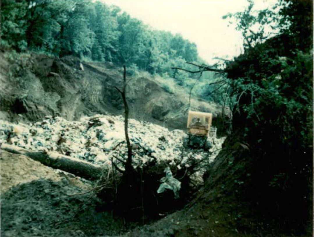 Trash and miscellany fill an open pit in the ground. An uprooted tree stump sits on its side in the foreground with trash embedded in the dirt-caked roots. A tractor plows over the trash pile in the background. 