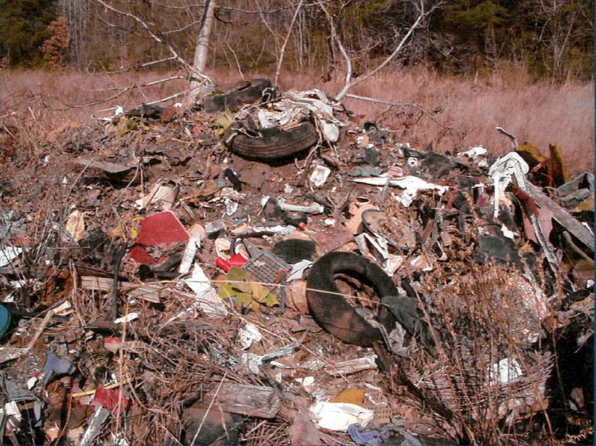 An uncovered debris pile of automobile shredding residue containing dirt, tires, shards of metal, and seat fluff; surrounded by trees and grass.