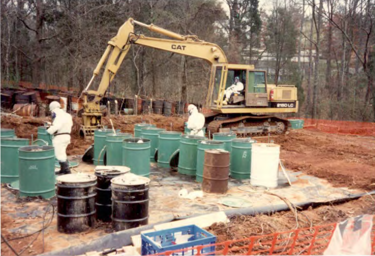 Cleanup of dozens of green and brown steel drums is conducted by workers in white haz-mat suits.  