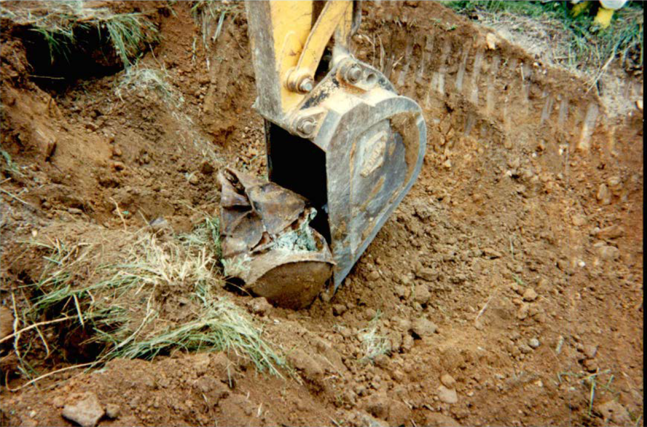 An excavator bulldozer rests the digging arm on a crushed rusty metal drum containing a strange greenish oxide in a large dirt pit.