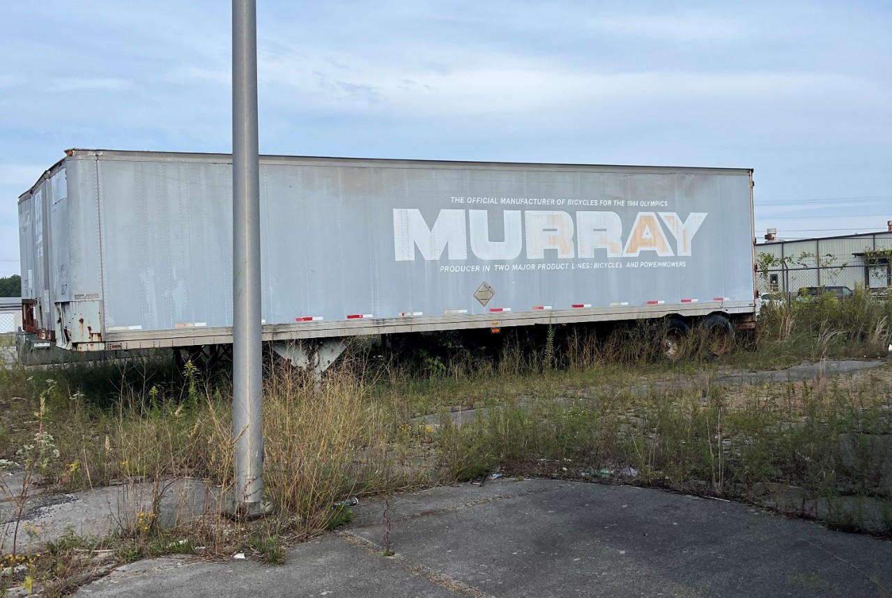 A grassy parking lot with an old truck with faded and bleached decal reading "The official manufacturer of bicycles for the 1984 Olympics: Murray: producer in two major product lines: bicycles and powermowers." 