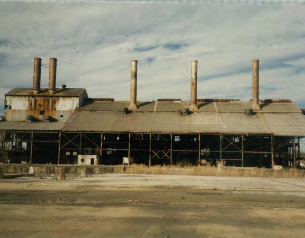 The since-dempolished Chromasco facility furnace building featuring rusty roof and iron scaffolding below an ominous smokestack on a yellow-grassy concrete slab..