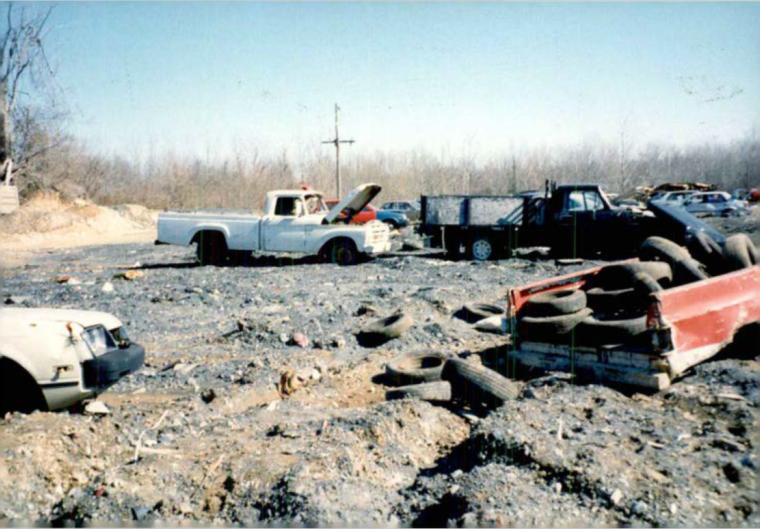 Auto salvage yard on the Fiberfine site. A gray, lumpy, topographically uneven gravel yard hosts sagging cars. A white 1980s Chevy truck in the background with its hood parked. Old tires spill out of a red truckbed in the foreground.